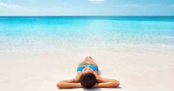 Tropical Caribbean beach vacation - suntan relaxation woman. Bikini girl lying down relaxing on white sand in the Bahamas. Girl sunbathing during summer holidays or winter getaway.