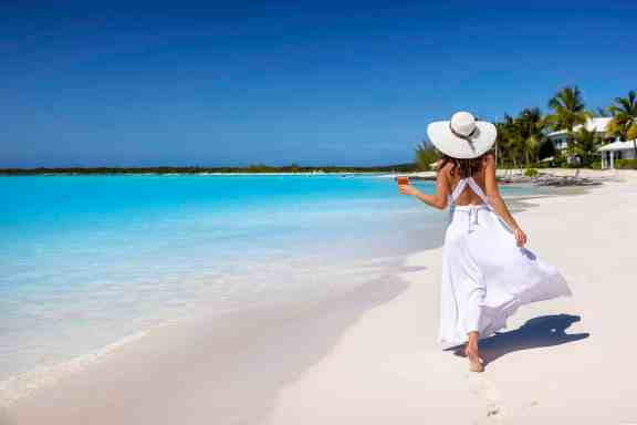 A tourist woman in a white summer dress and holding a drink walks along a tropical beach with turquoise sea and palm trees