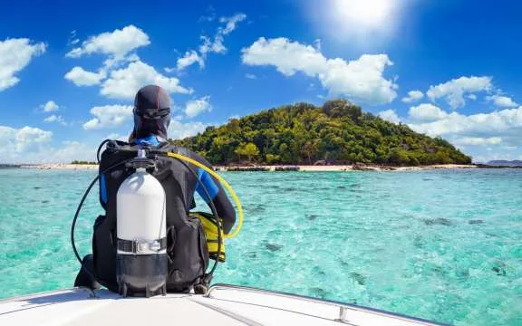 A scuba diver in his diving gear sits in front of a boat and enjoys the view of the tropical landscape with turquoise sea