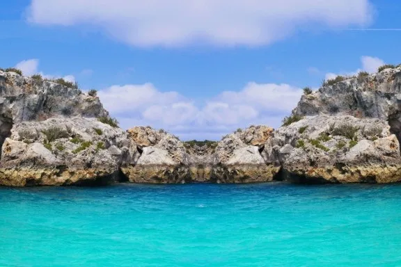 This image showcases the striking entrance to Thunderball Grotto in the Bahamas, an iconic underwater cave known for its use in the James Bond film "Thunderball." The grotto's rugged limestone cliffs, topped with sparse vegetation, frame the turquoise waters that lead into the cavern. The symmetrical composition highlights the natural beauty and mystery of the site, inviting visitors to explore what lies beneath the surface in this popular snorkeling and diving location.