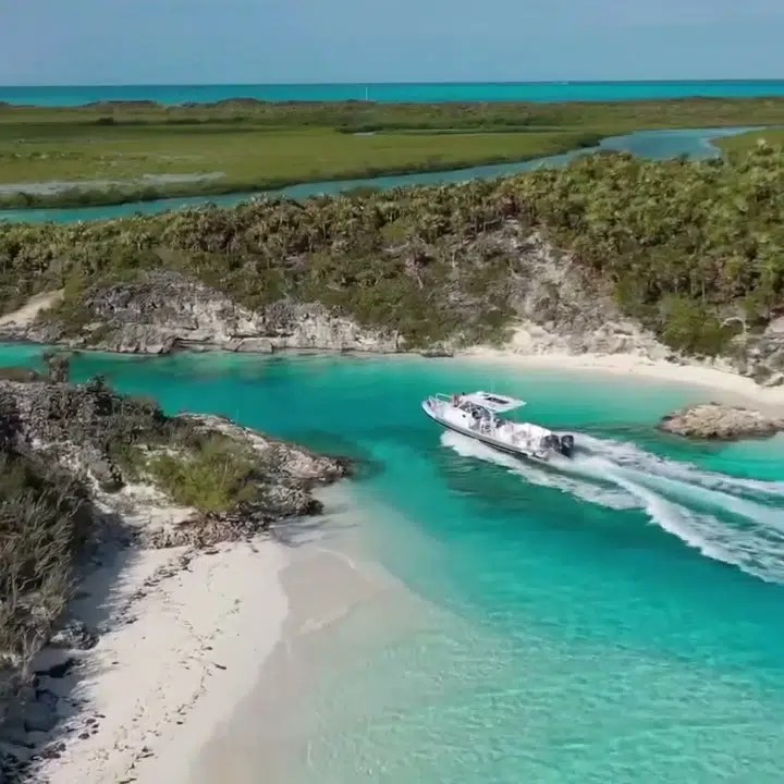 A tender curves through a clear turquoise channel between white sand and low Bahamas cays